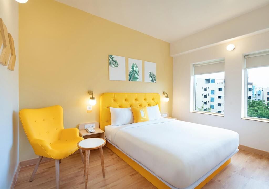 Een hotelkamer met een bed, een gele fauteuil, een klein tafeltje en een nachtkastje. Er zijn drie wandlampen en een raam met uitzicht op gebouwen.