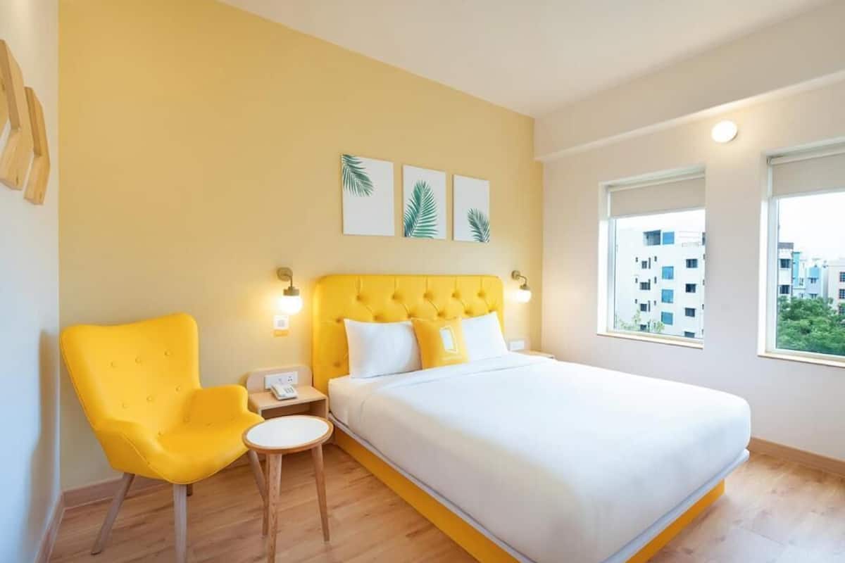 A hotel room with a bed, a yellow armchair, a small table, and a nightstand. There are three wall-mounted light fixtures and a window with a view of buildings.