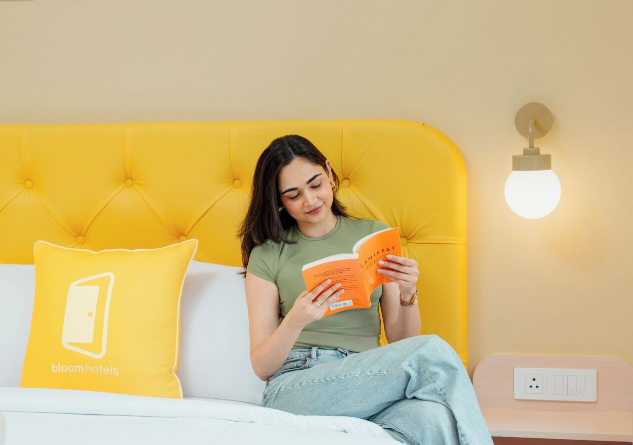 Iemand zit op een bed met een geel hoofdeinde, en leest een boek. Er ligt een kussen met een logo op, een nachtkastje met een lamp, en een stopcontact.