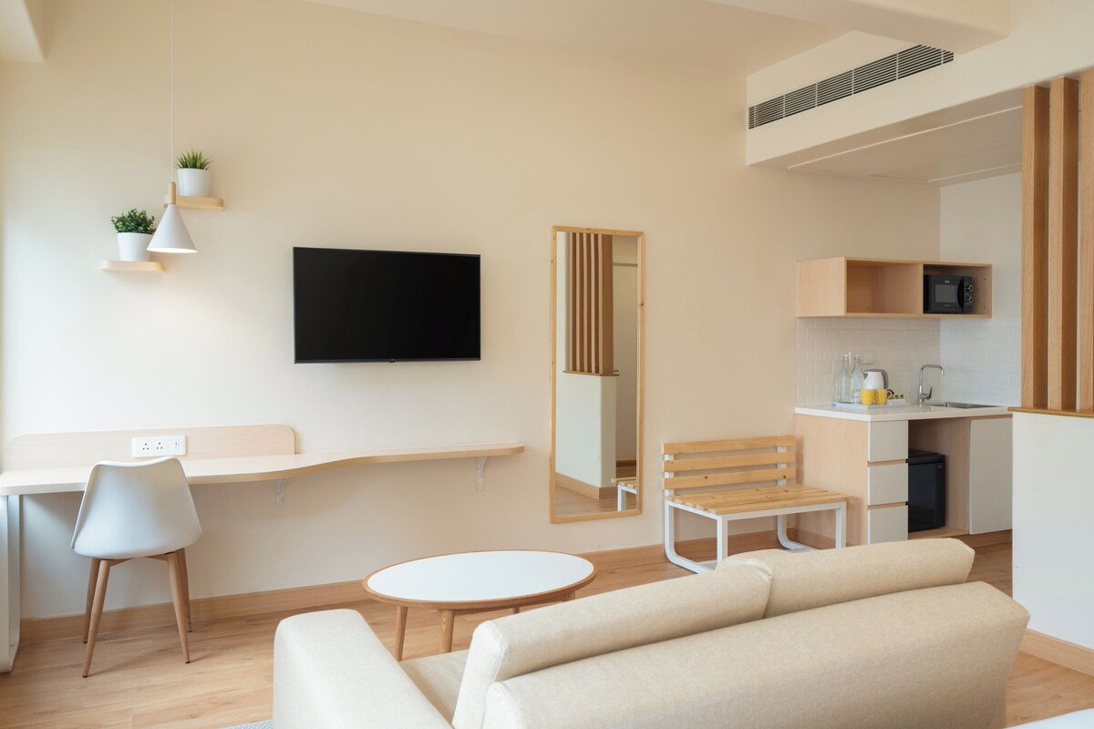 Una habitación de hotel moderna con televisor de pantalla plana, un escritorio con silla, un sofá, una mesita redonda y una cocina equipada con microondas y fregadero.