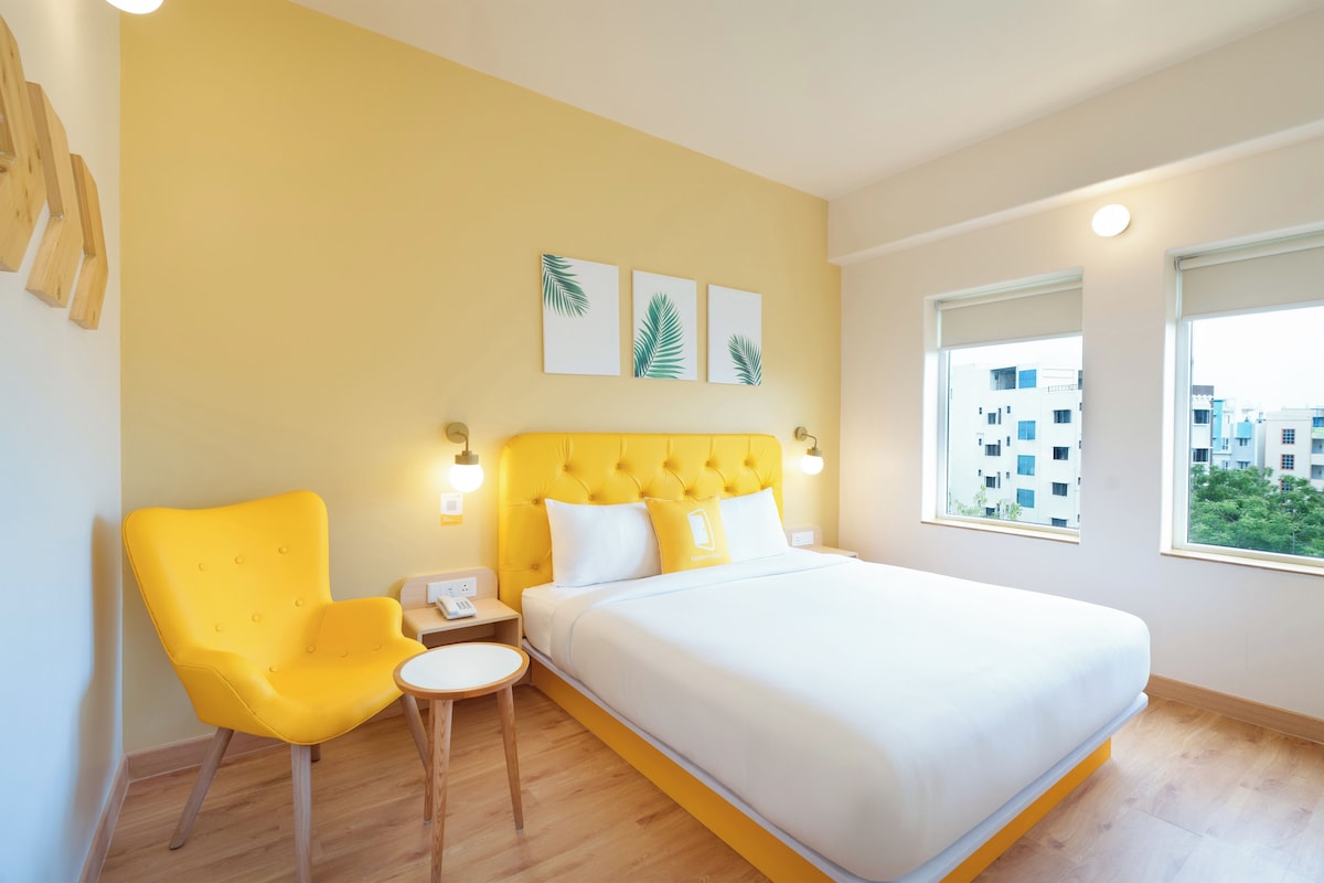A hotel room with a bed, a chair, a bedside table, and a window with a view of buildings.
