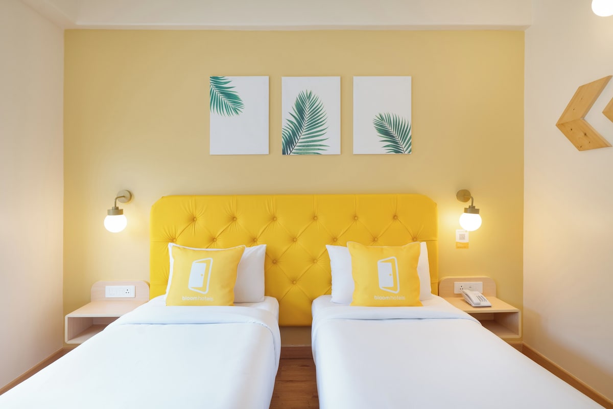 Habitación de hotel con cabecera amarilla, dos camas, ropa de cama blanca y tres adornos de hojas colgados en la pared.
