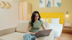 Una persona sentada en un sofá con una computadora portátil, una cama con cabecera amarilla y una mesita de noche con una lámpara en una habitación con piso de madera.