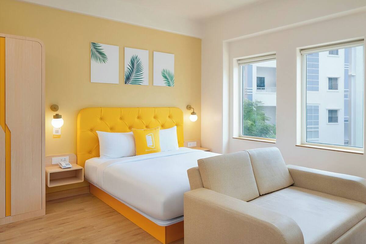 A hotel room with a yellow bed, white bedding, a beige sofa, and a window with a view of buildings.