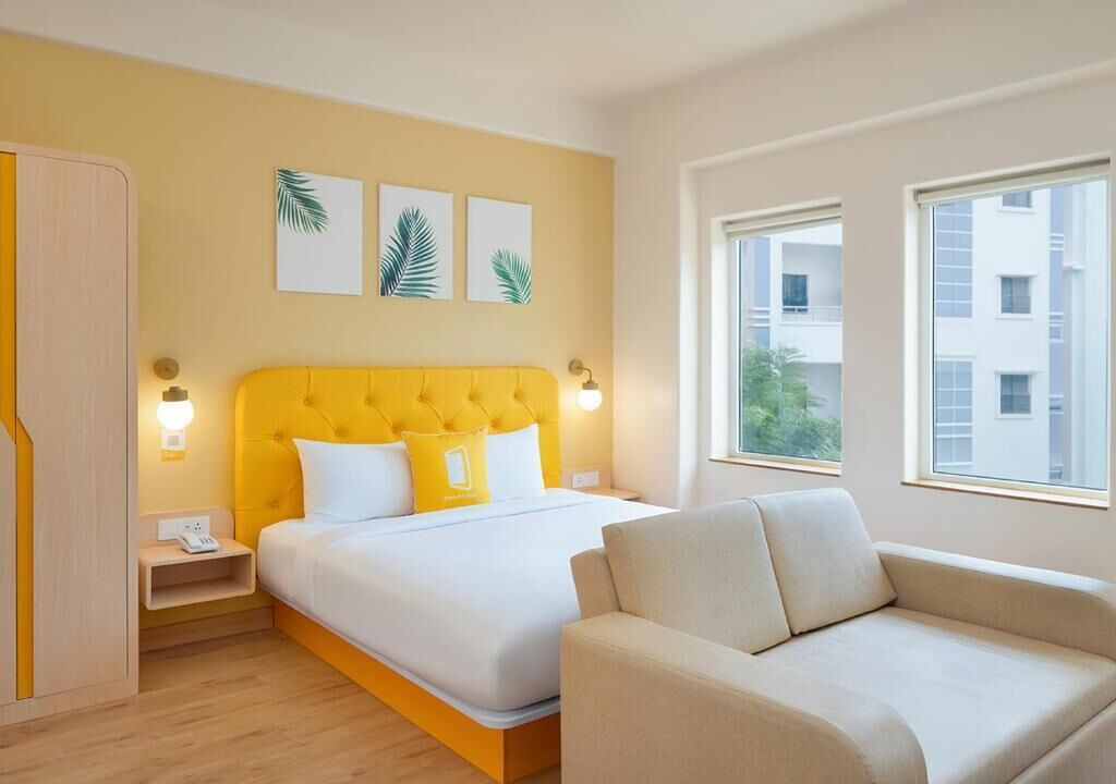 Een hotelkamer met een geel hoofdeinde, een bed met wit beddengoed, een beige bank en een raam met uitzicht op gebouwen.