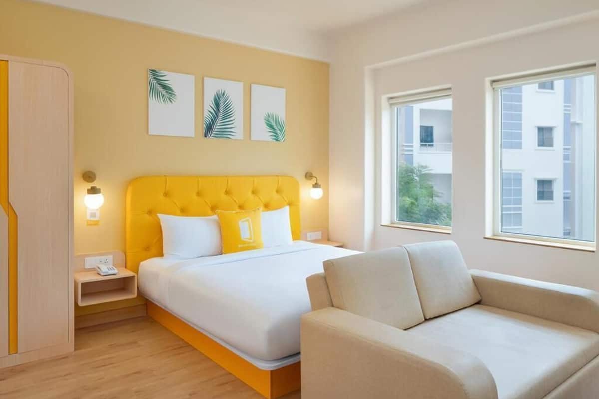A hotel room with a yellow headboard, a bed with white bedding, a beige sofa, and a window with a view of buildings.