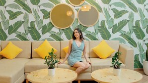 Una mujer sentada en un sofá beige con almohadas amarillas, rodeada por dos mesas redondas de madera con plantas en macetas.