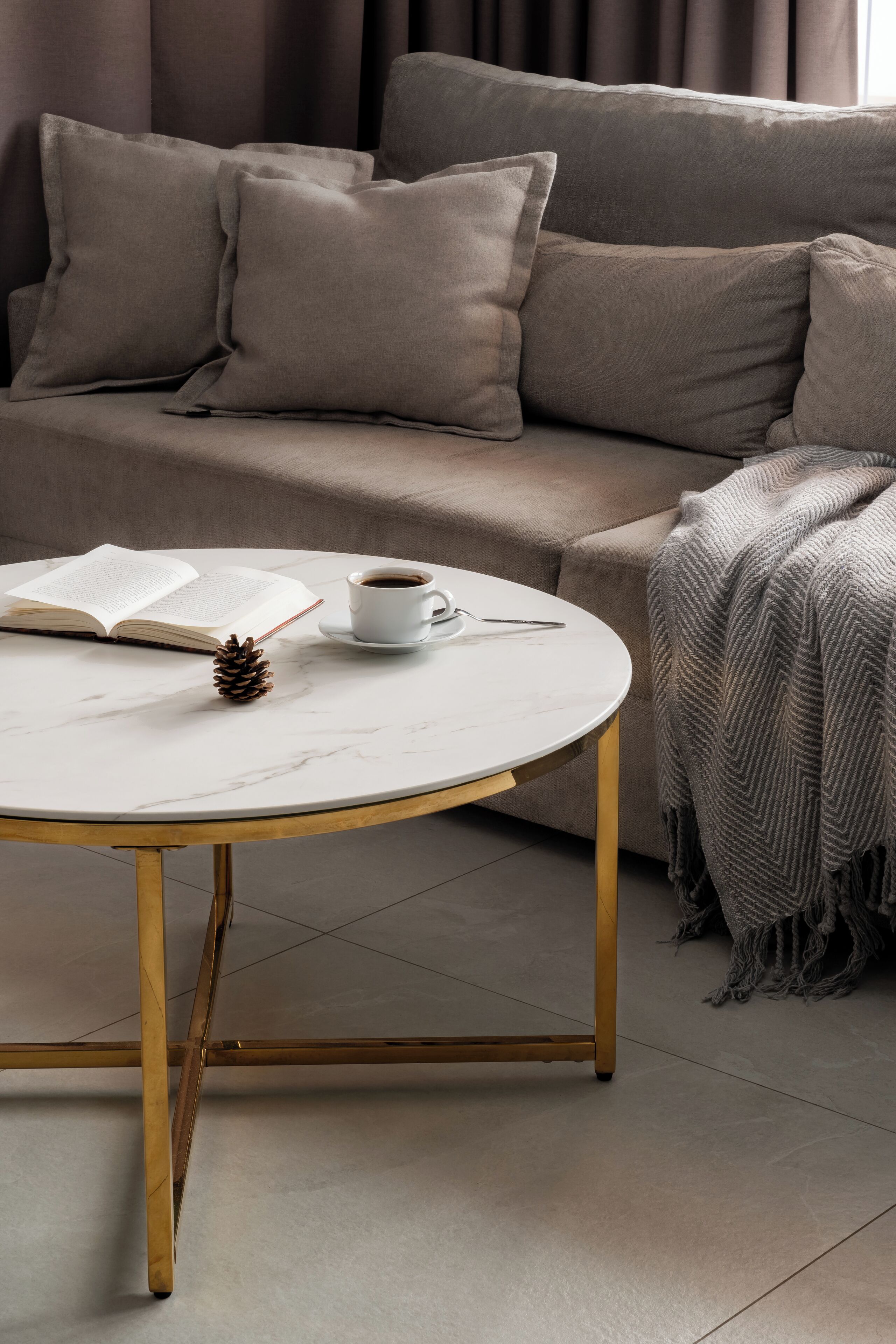 A round white table with a gold base, a book, a coffee cup, and a pine cone on it.