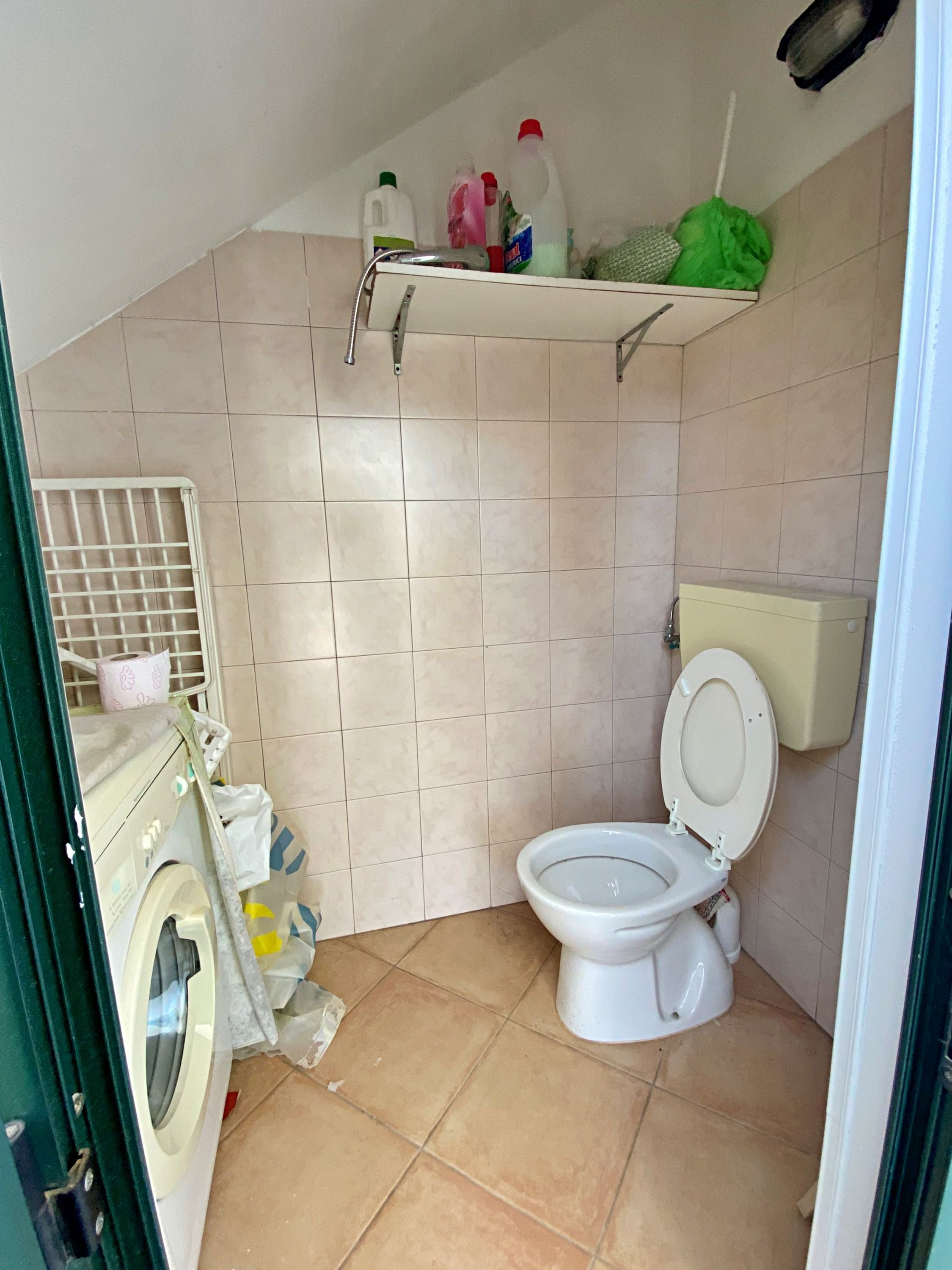 A small, tiled bathroom with a toilet, a washing machine, and a shelf with cleaning supplies.