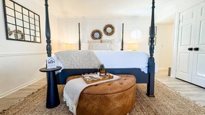 A bedroom with a four-poster bed, a bedside table with a WiFi sign, a brown ottoman, and a decorative mirror on the wall.