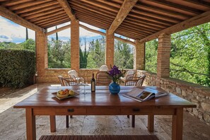 Une terrasse couverte avec une table en bois, des chaises, un vase rempli de fleurs, une bouteille et un saladier de fruits.