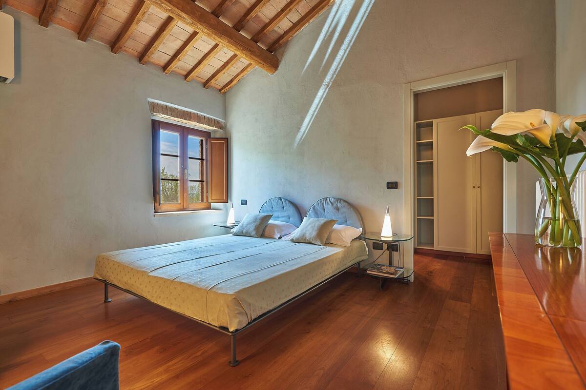 Une chambre à coucher avec un lit, un plancher en bois, une fenêtre, une table de chevet avec une lampe et un vase de fleurs.