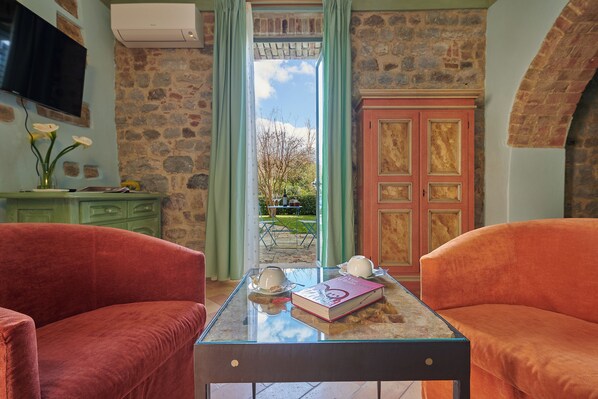 Una habitación con pared de piedra, una mesa de vidrio con un libro y tazas de té, dos sofás rojos y vistas a través de una puerta.