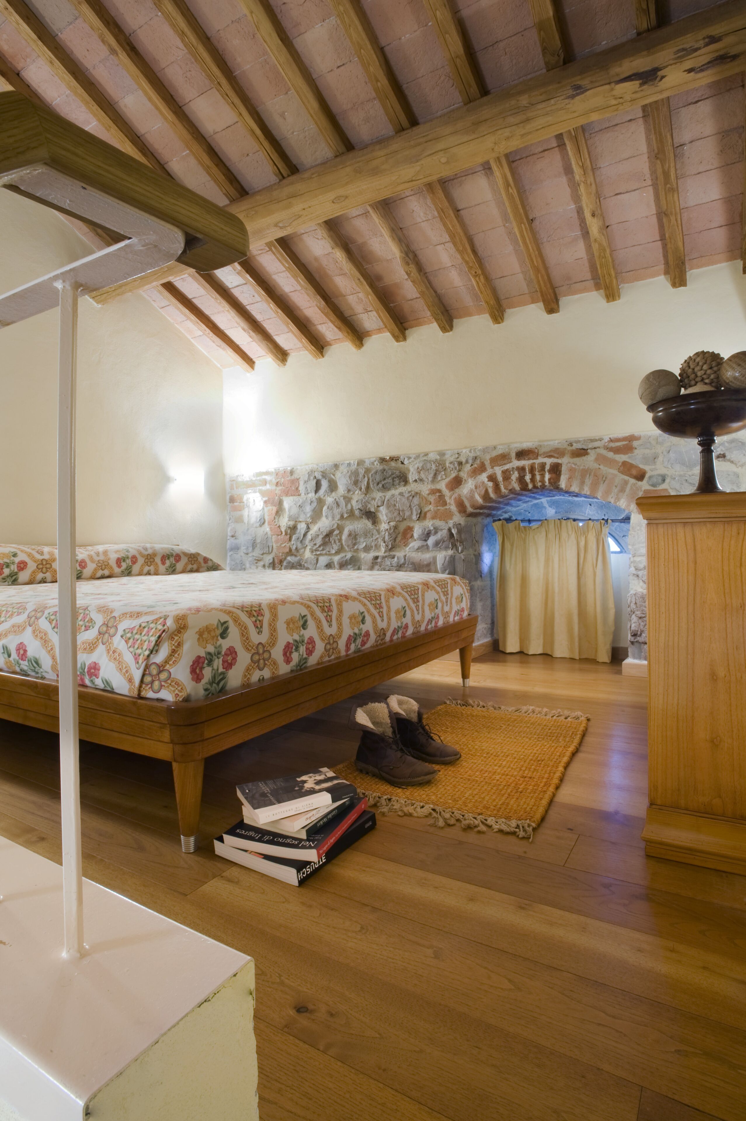 A bedroom with a wooden ceiling, a stone wall, a bed with patterned bedding, a bedside table, and a nightstand.