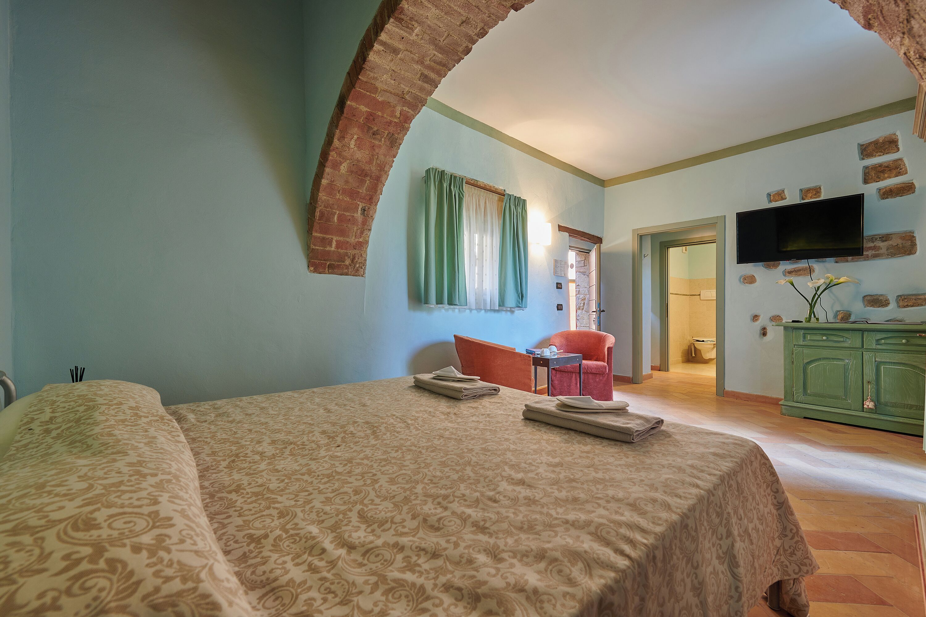 Quarto de hotel com uma cama, televisão, um armário verde e um arco de tijolos.
