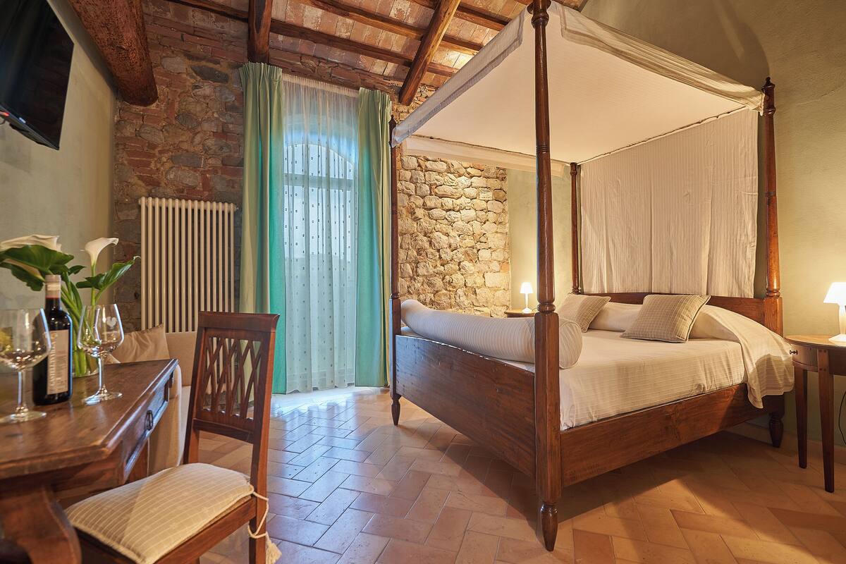 Un lit à baldaquin avec un mur en pierre en arrière-plan, une table en bois avec des verres à vin et une chaise avec un coussin.