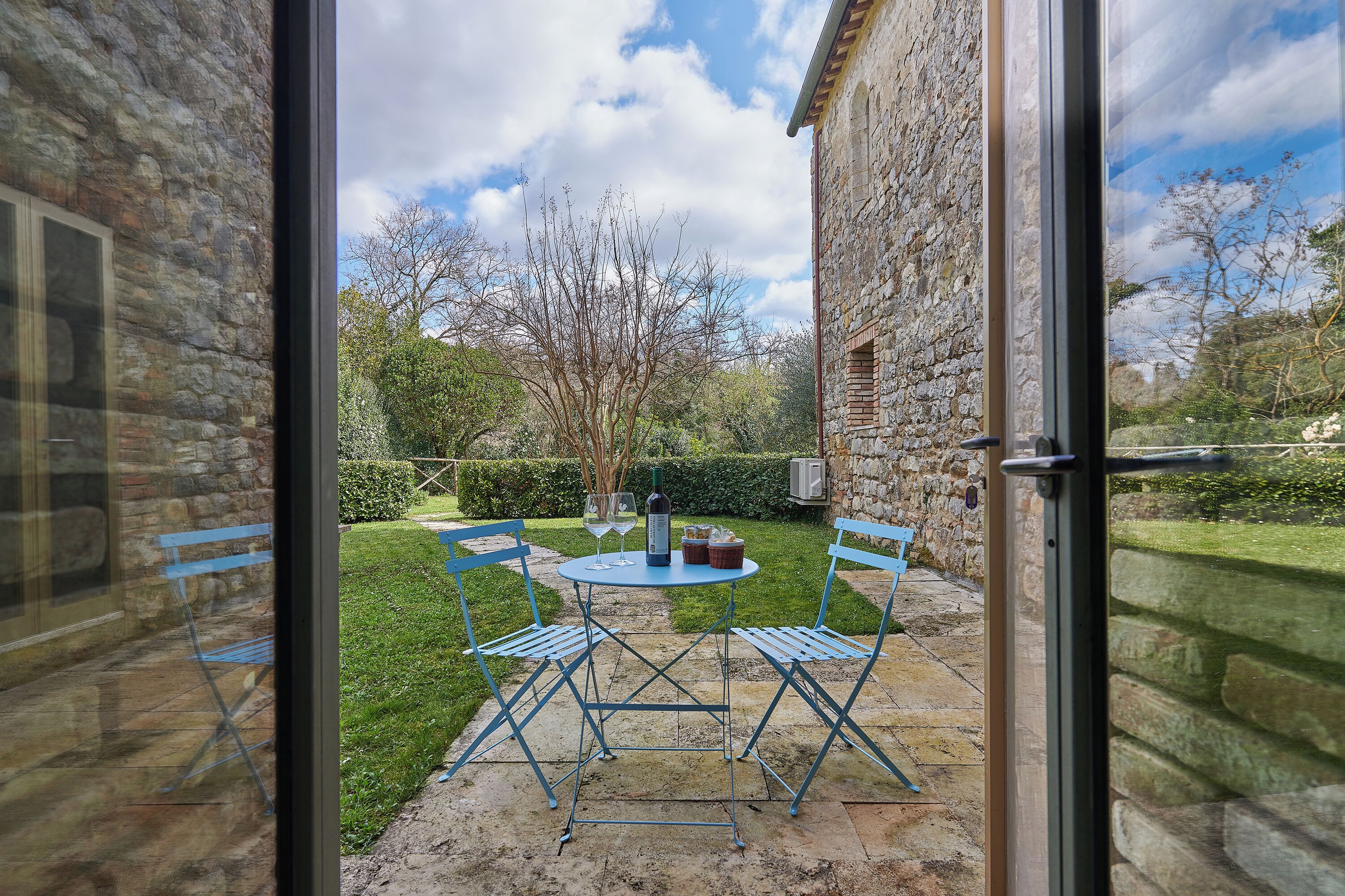A stone building with a glass door, a small patio with blue chairs and a table, and a grassy garden in the background.