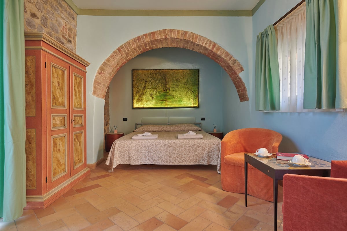Une chambre avec une arche en briques, un lit recouvert d’une couvre-lit à motifs, une table de chevet, une chaise et une table sur laquelle se trouvent un livre et une tasse.