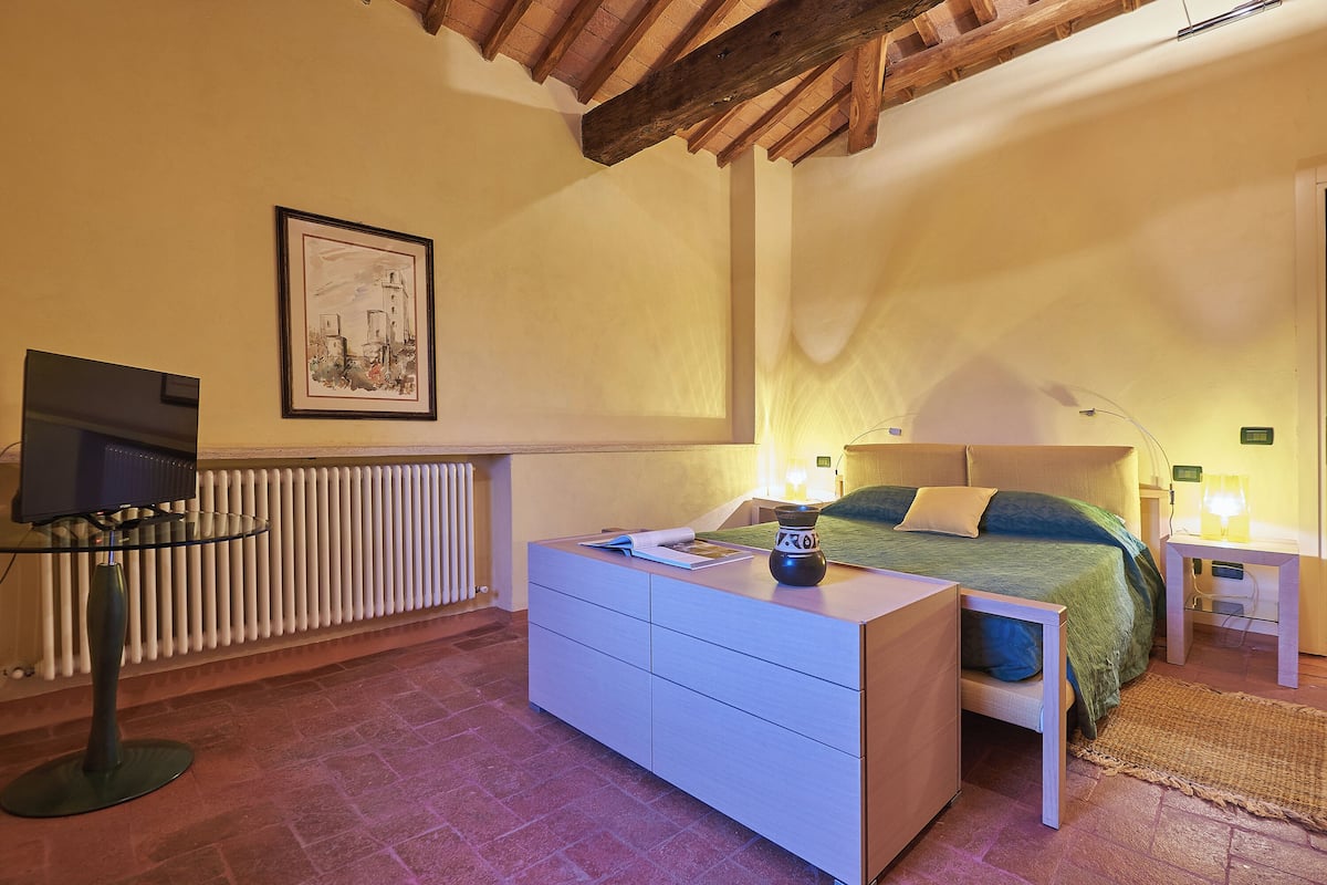 Une chambre à coucher comprenant un lit, des tables de chevet, une commode, un radiateur, un tableau encadré au mur et un téléviseur posé sur un meuble.