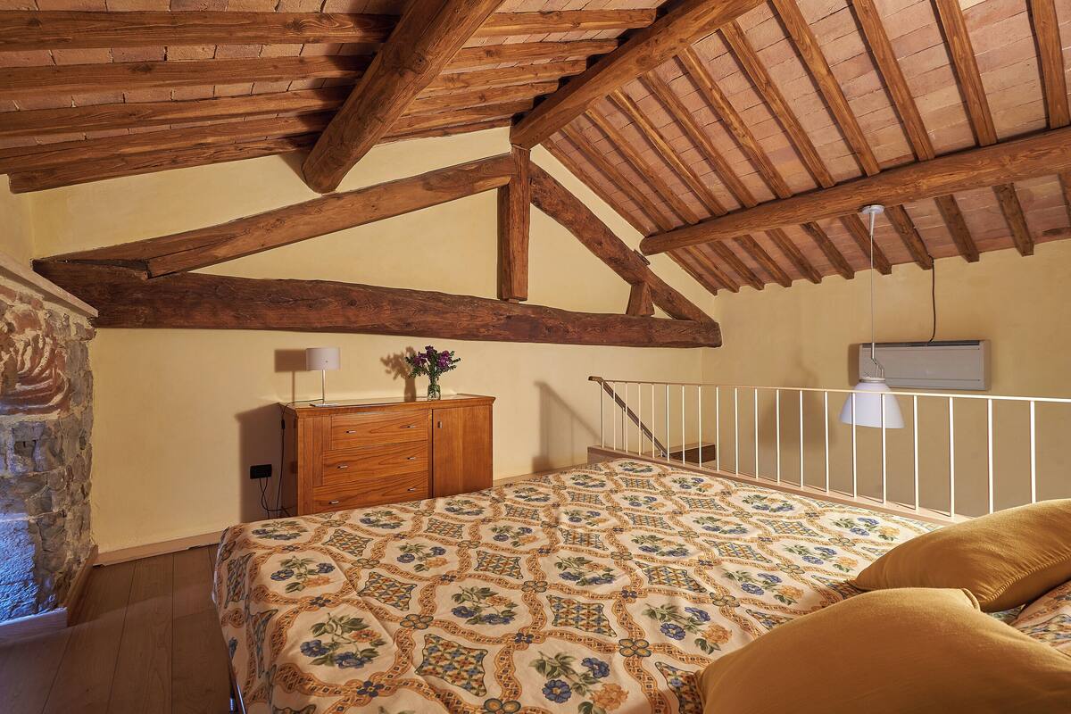 Une chambre avec un plafond en bois, un lit recouvert d’une couvre-lit à motifs, une commode et une balustrade blanche.