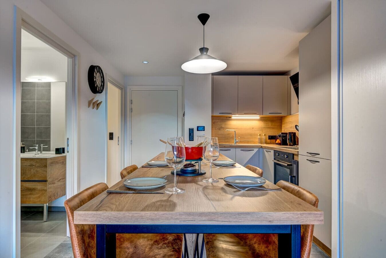 A modern kitchen with a wooden dining table, a hanging light fixture, and a clock on the wall.