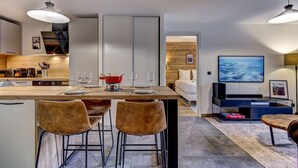 A modern kitchen with a dining area, a TV, and a bed in the background.