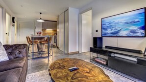 A modern living room with a large flat-screen TV, a comfortable sofa, a wooden coffee table, and a dining area with chairs and a kitchen in the background.