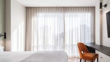 Une chambre d’hôtel avec un grand lit, un bureau et une chaise. Il y a une fenêtre avec des rideaux légers et une vue sur la ville.