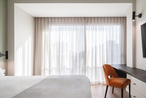 A hotel room with a large bed, a desk, and a chair. There is a window with sheer curtains and a view of the city. -  Limehome Lisbon Campo Pequeno (Lisbon)