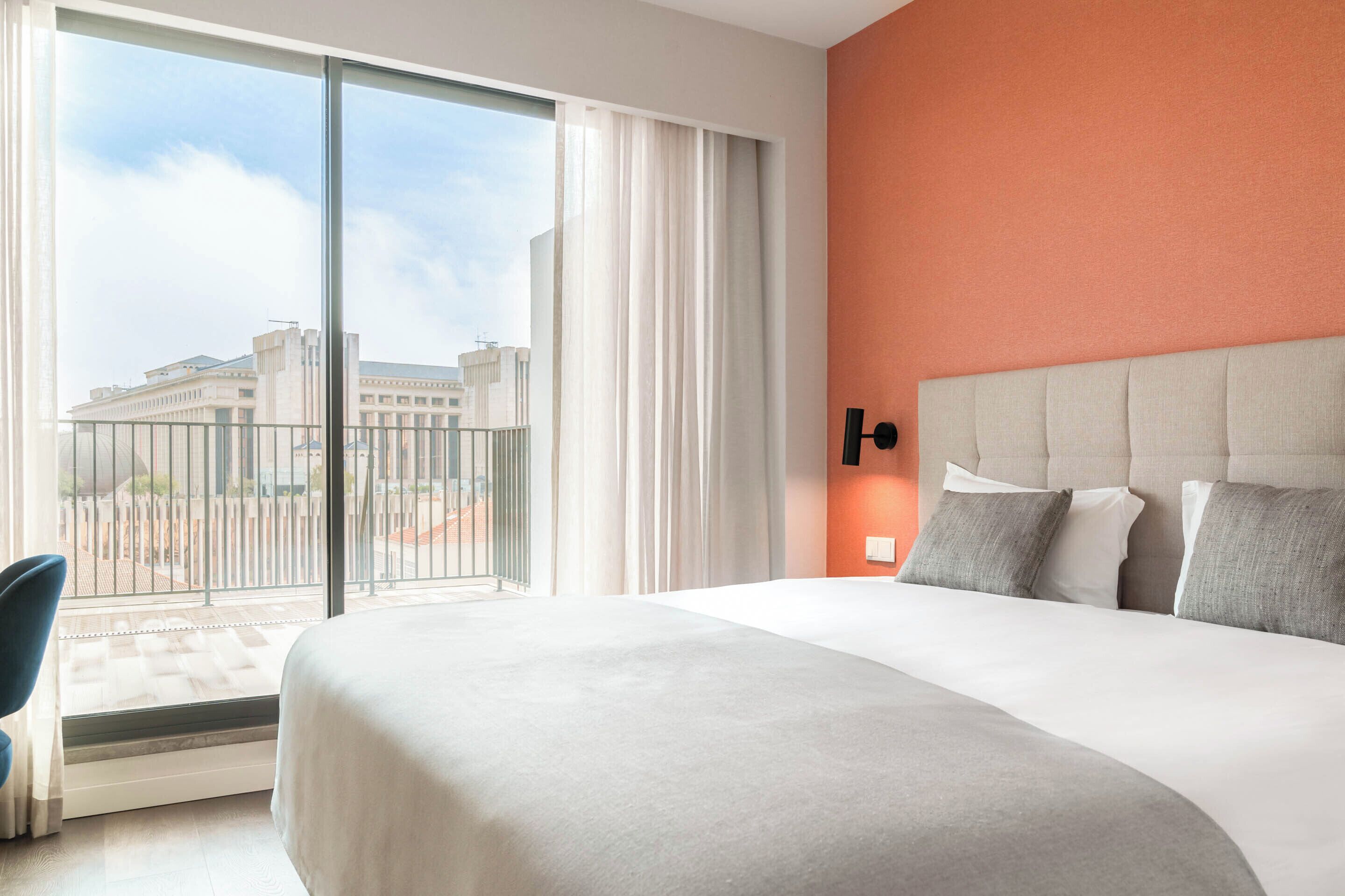Ein Hotelzimmer mit einem großen Bett, Blick auf ein Gebäude durch eine Glastür und einer roten Akzentwand.