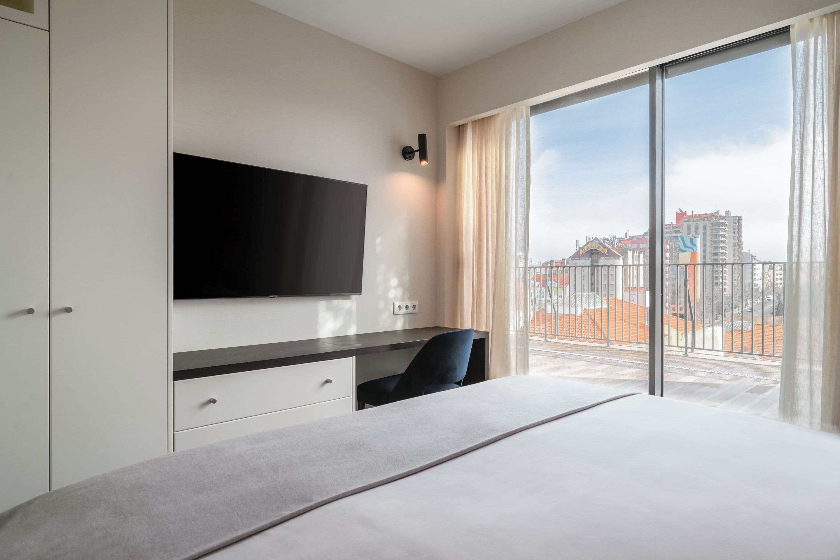 Ein Hotelzimmer mit einem grossen Bett, einem flachbildfernseher an der Wand, einem Schreibtisch und Blick auf die Stadt durch grosse Fenster.