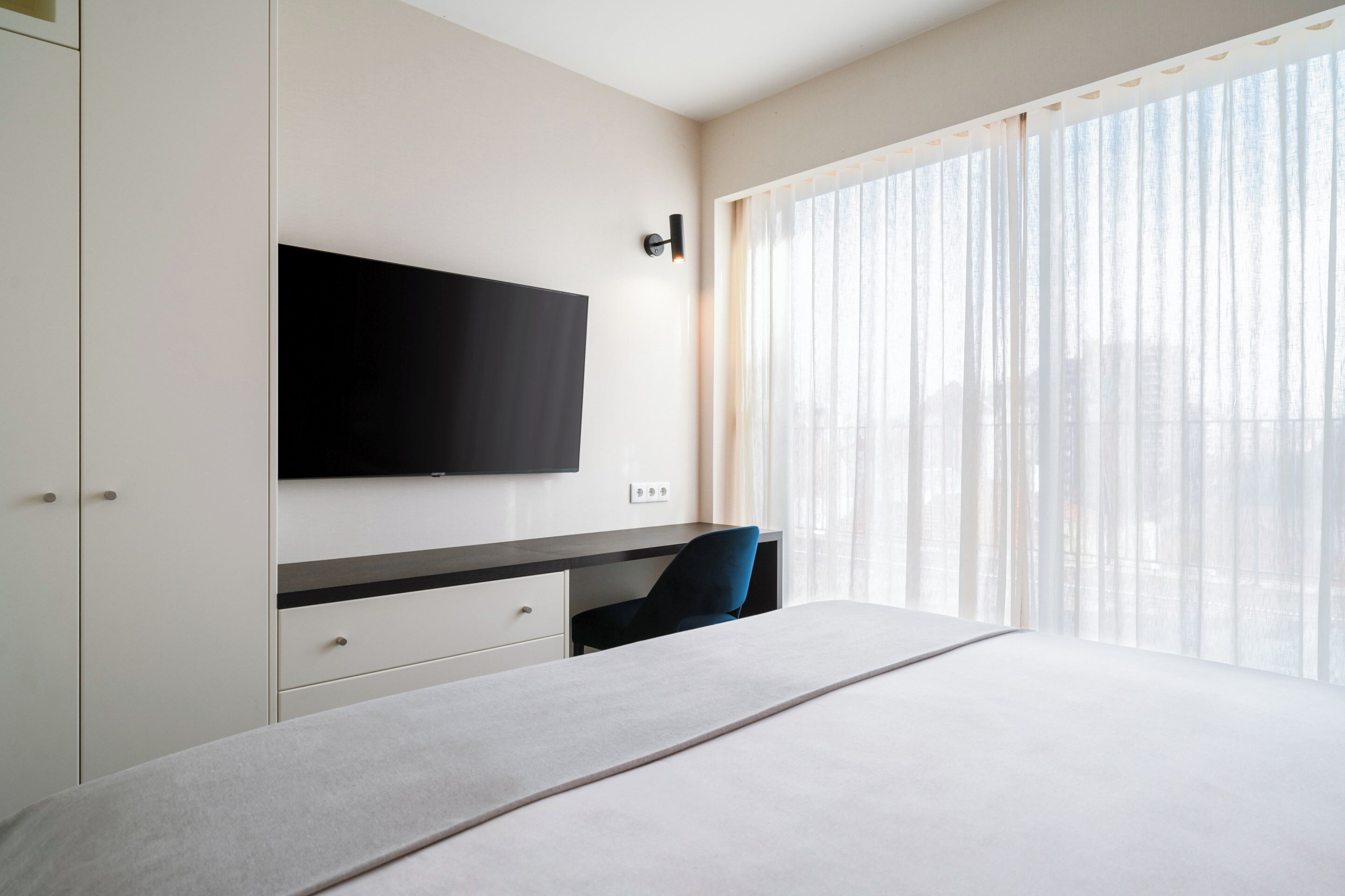Una habitación de hotel moderna con una cama grande, un televisor de pantalla plana montado en la pared, un escritorio con una silla y un amplio ventanal con cortinas de encaje.
