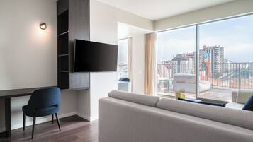 A modern living room with a flat-screen TV mounted on the wall, a sofa, a chair, and a large window offering a view of the cityscape.