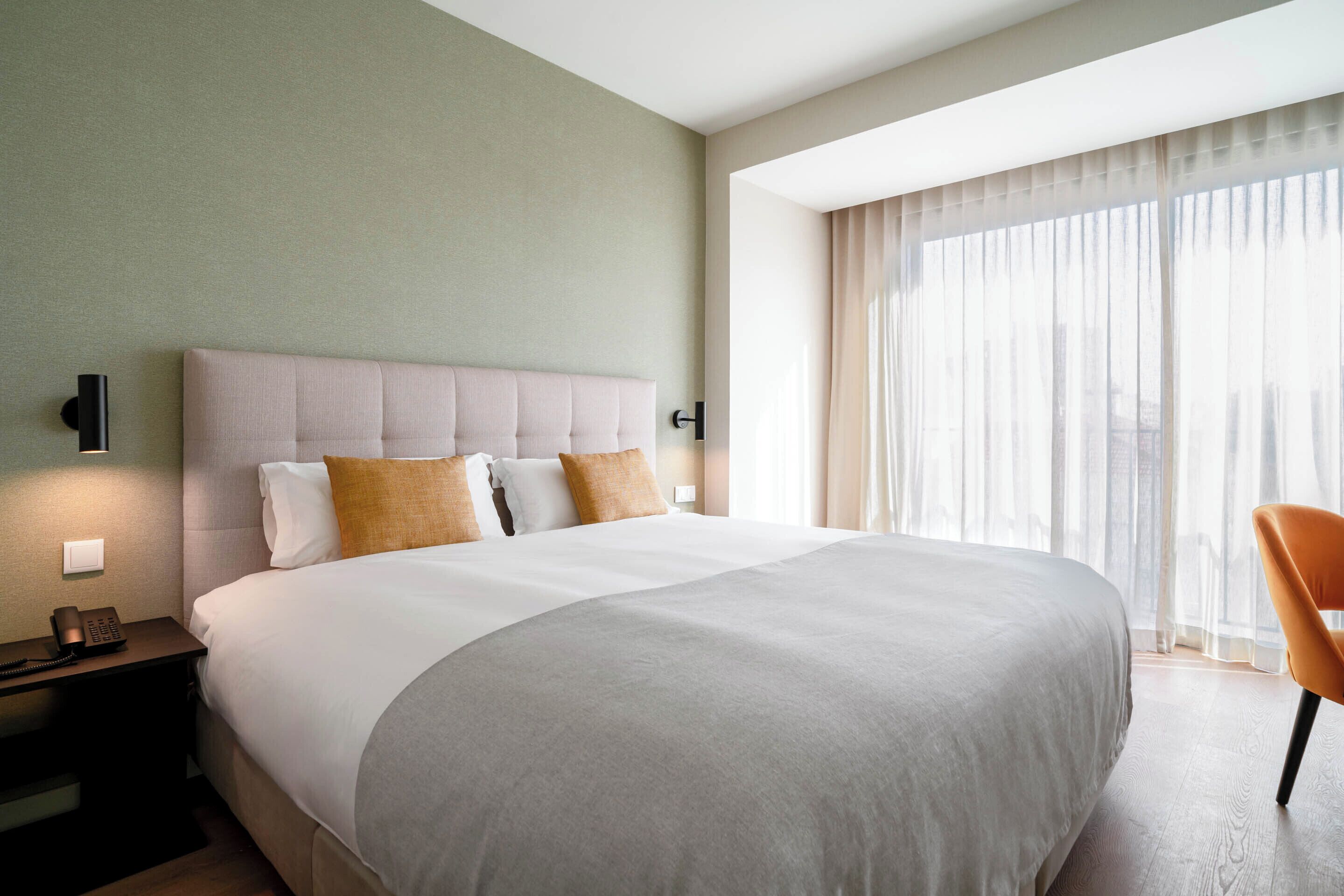 A hotel room with a large bed, a bedside telephone, and a chair near a window with sheer curtains.