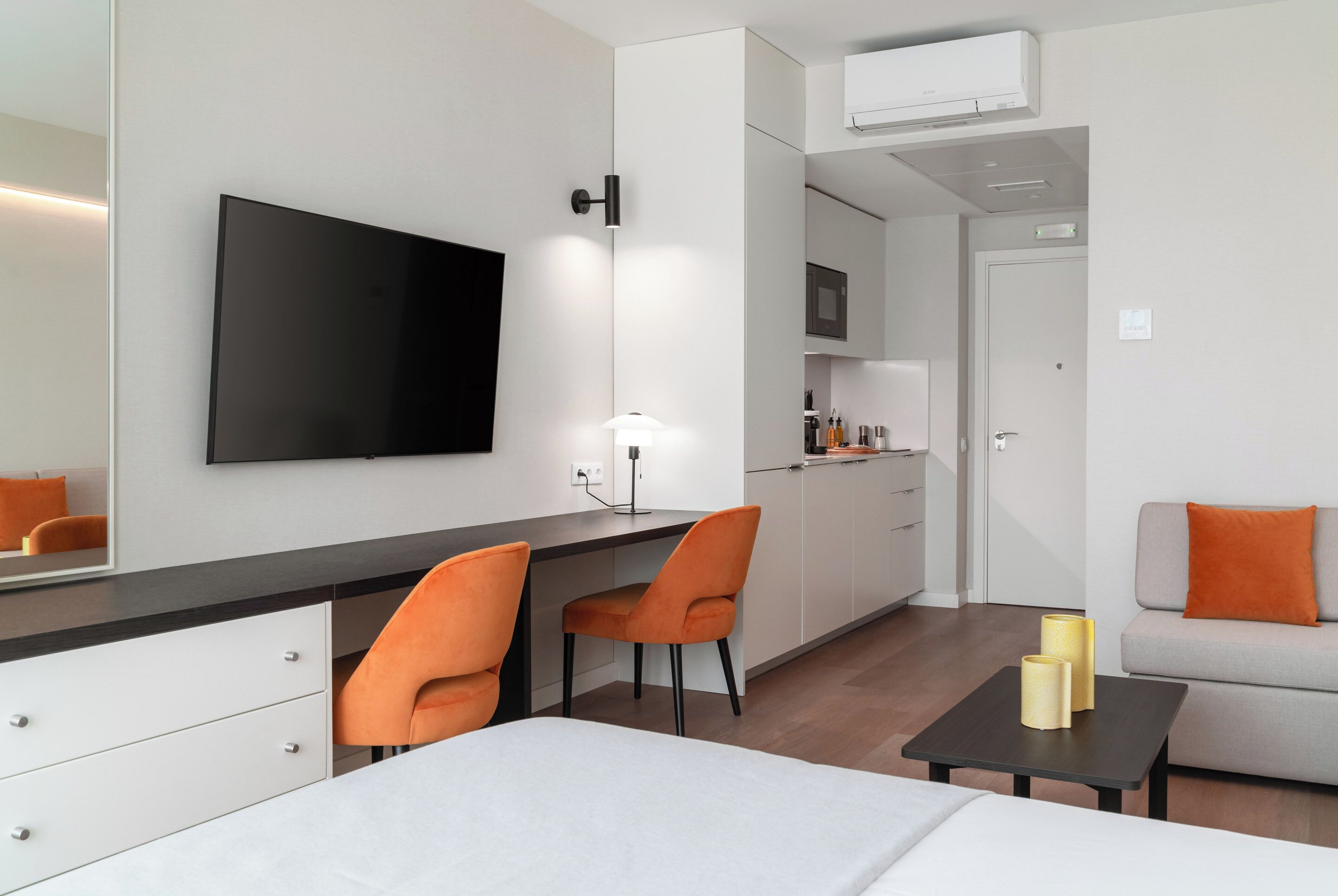 A modern hotel room with a bed, a desk with two chairs, a TV, a small table with two candles, and a sofa with an orange pillow.