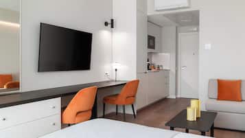 A modern hotel room with a bed, a desk with two chairs, a TV, a small table with two candles, and a sofa with an orange pillow.