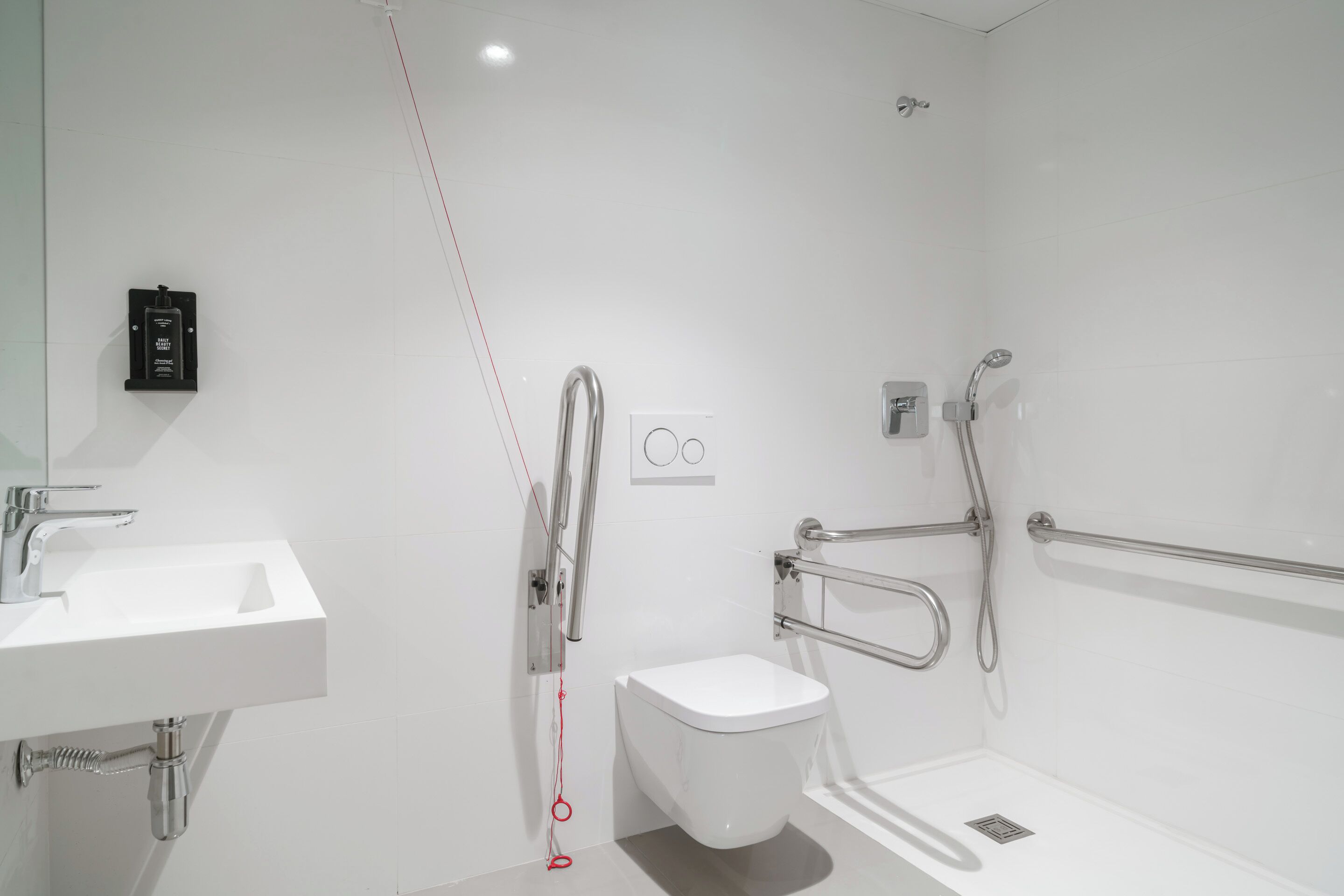 Ein modernes Badezimmer mit einem weißen Waschbecken, einer Toilette und einer Duschkabine, ausgestattet mit Haltegriffen und einer Sicherungsleine.