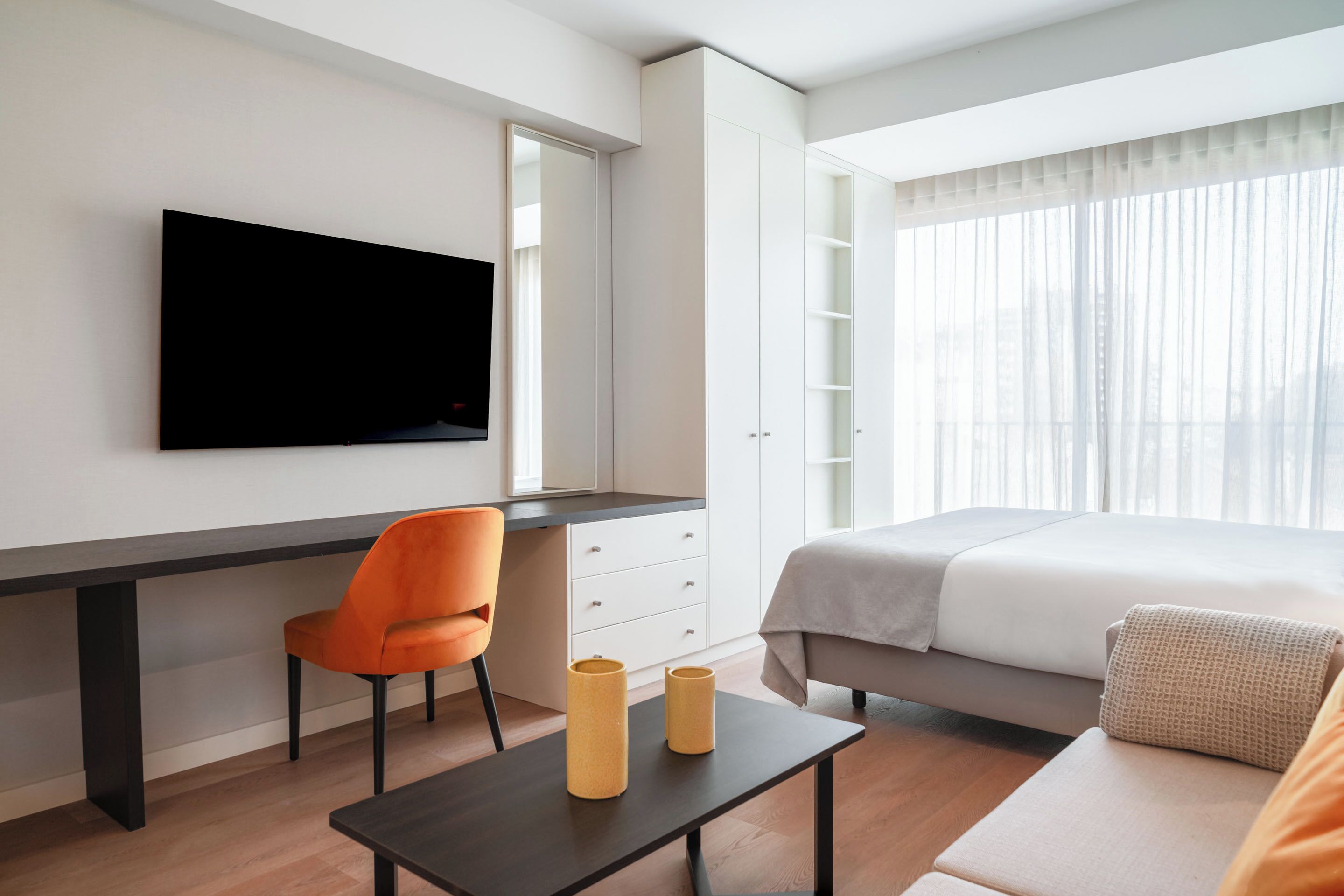 Quarto de hotel moderno com TV de tela plana na parede, uma escrivaninha com cadeira, uma cama com edredom branco e uma janela com cortinas translúcidas.