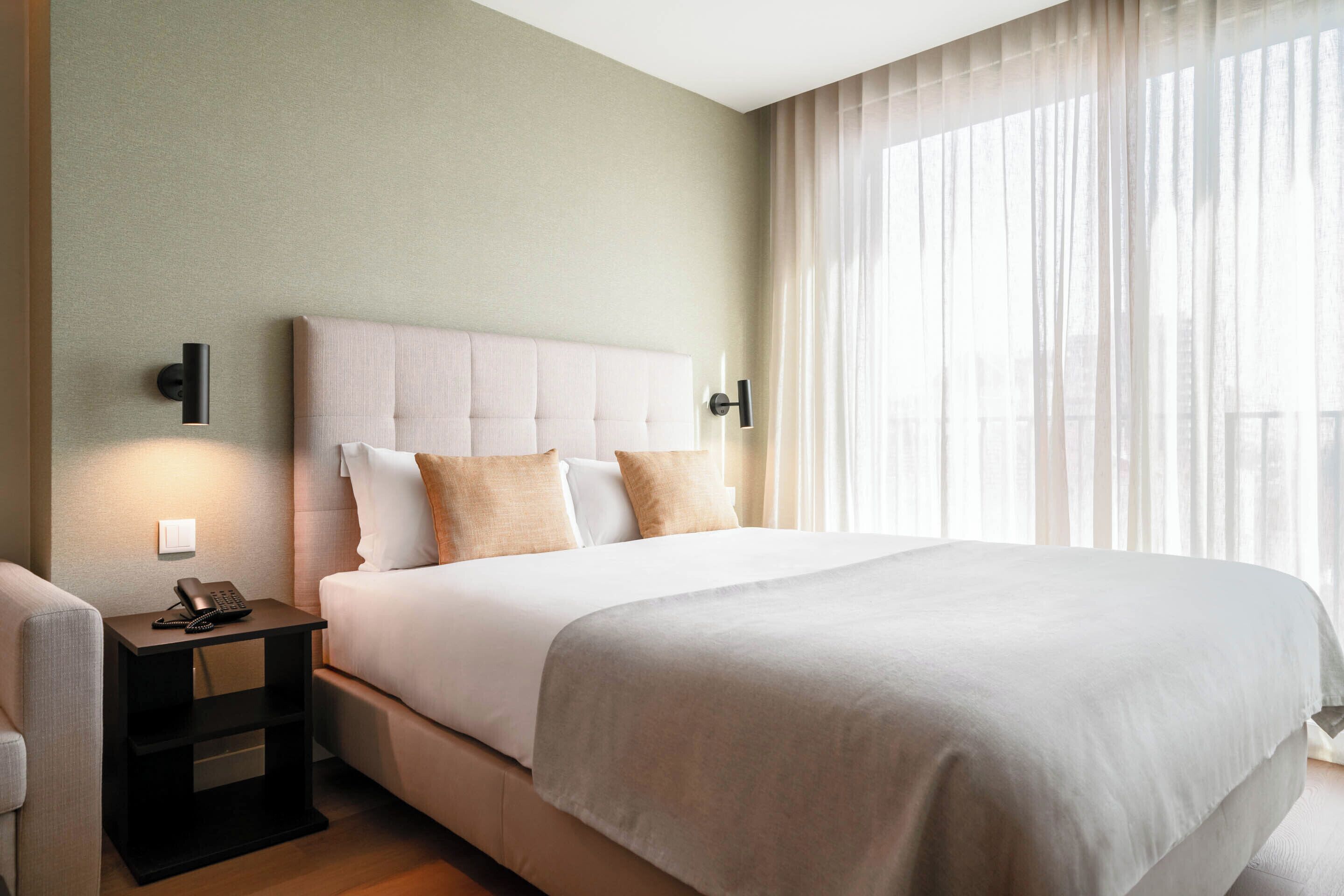 A hotel room with a large bed, a bedside table with a phone, and a view of the outdoors through sheer curtains.
