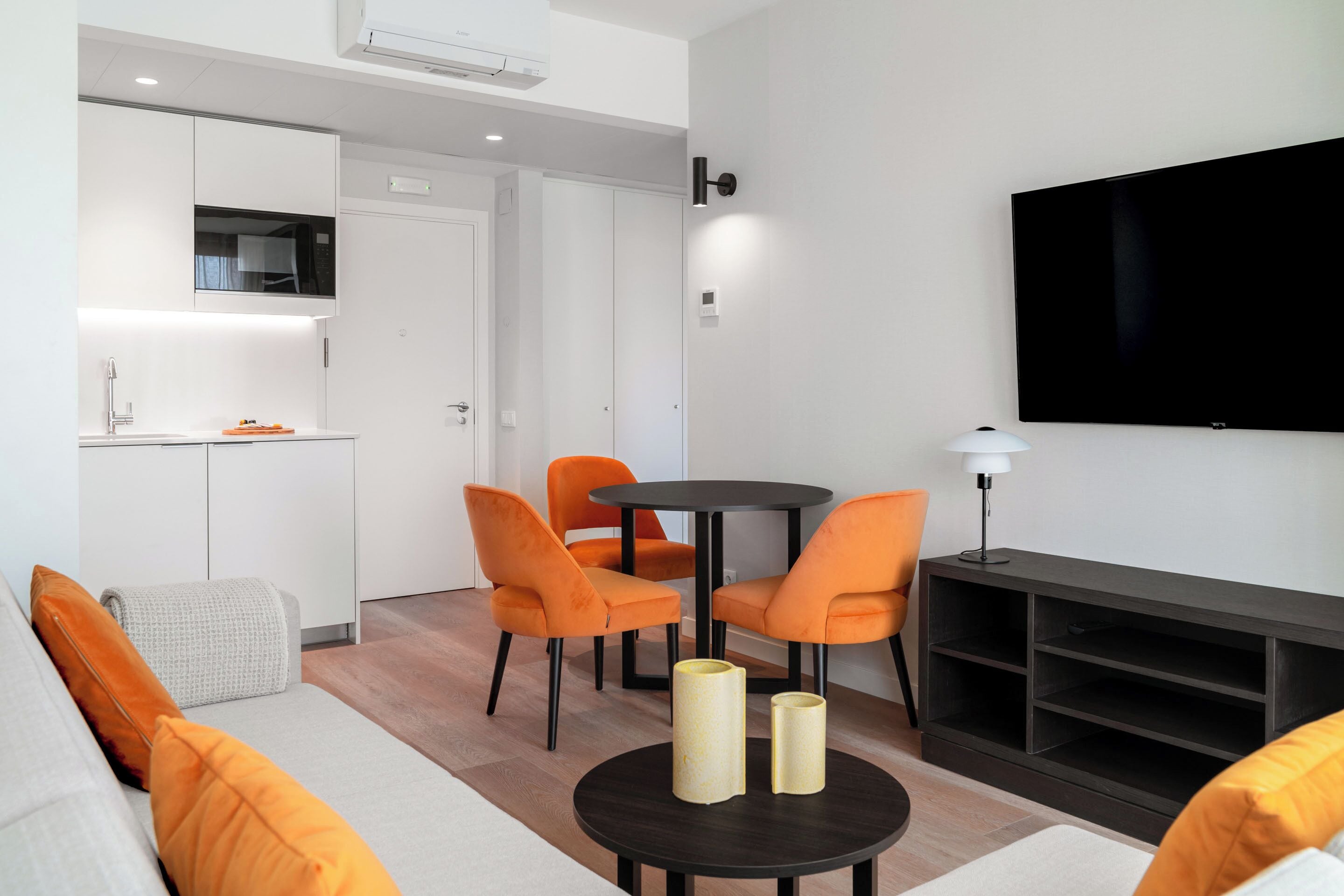 A modern living space with a sofa, orange chairs, a round table with candles, a TV, and a kitchen area.