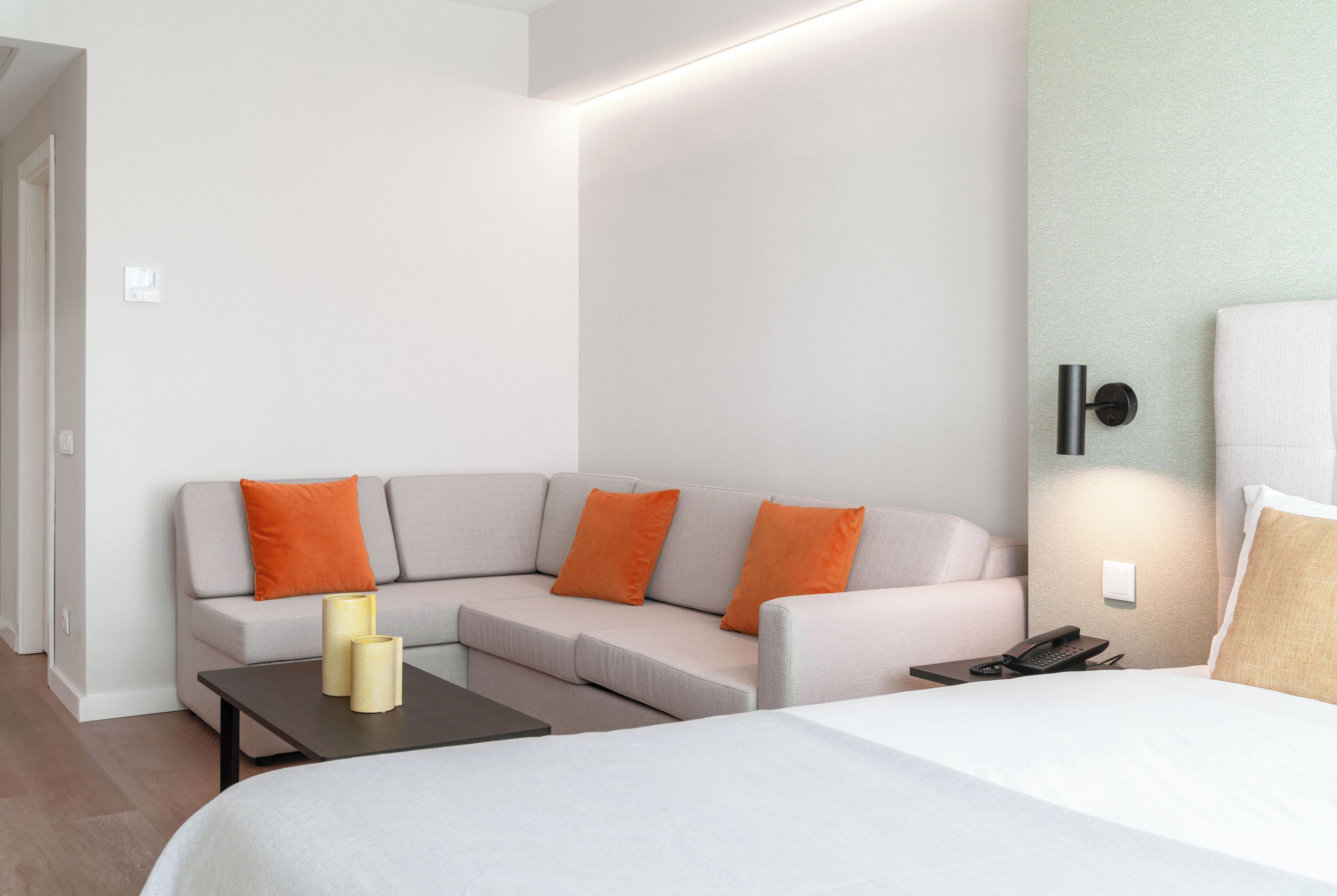 A modern hotel room with a large bed, a sofa with orange cushions, a small table with a lamp, and a bedside table with a phone.
