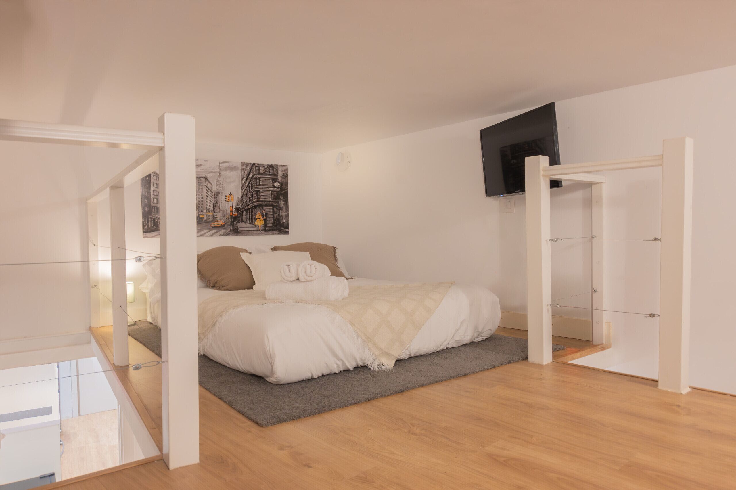 A bedroom with a bed, a gray rug, a painting on the wall, and a TV mounted on the wall.