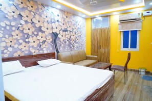 A hotel room with a large bed, a sofa, a wooden headboard, a wall mural, an air conditioning unit, and a window with blue shutters. - KAUSHIKI GARDEN (Patna)
