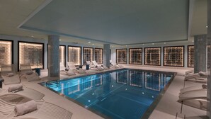 Una amplia piscina cubierta con sillas y camas para tomar el sol, rodeada de grandes ventanales con estantes iluminados.