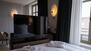 Habitación de hotel con una televisión de pantalla plana grande, una cama con sábanas blancas, dos toallas enrolladas, una silla y una ventana con cortinas.