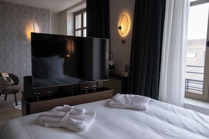 Habitación de hotel con una televisión de pantalla plana grande, una cama con sábanas blancas, dos toallas enrolladas, una silla y una ventana con cortinas.