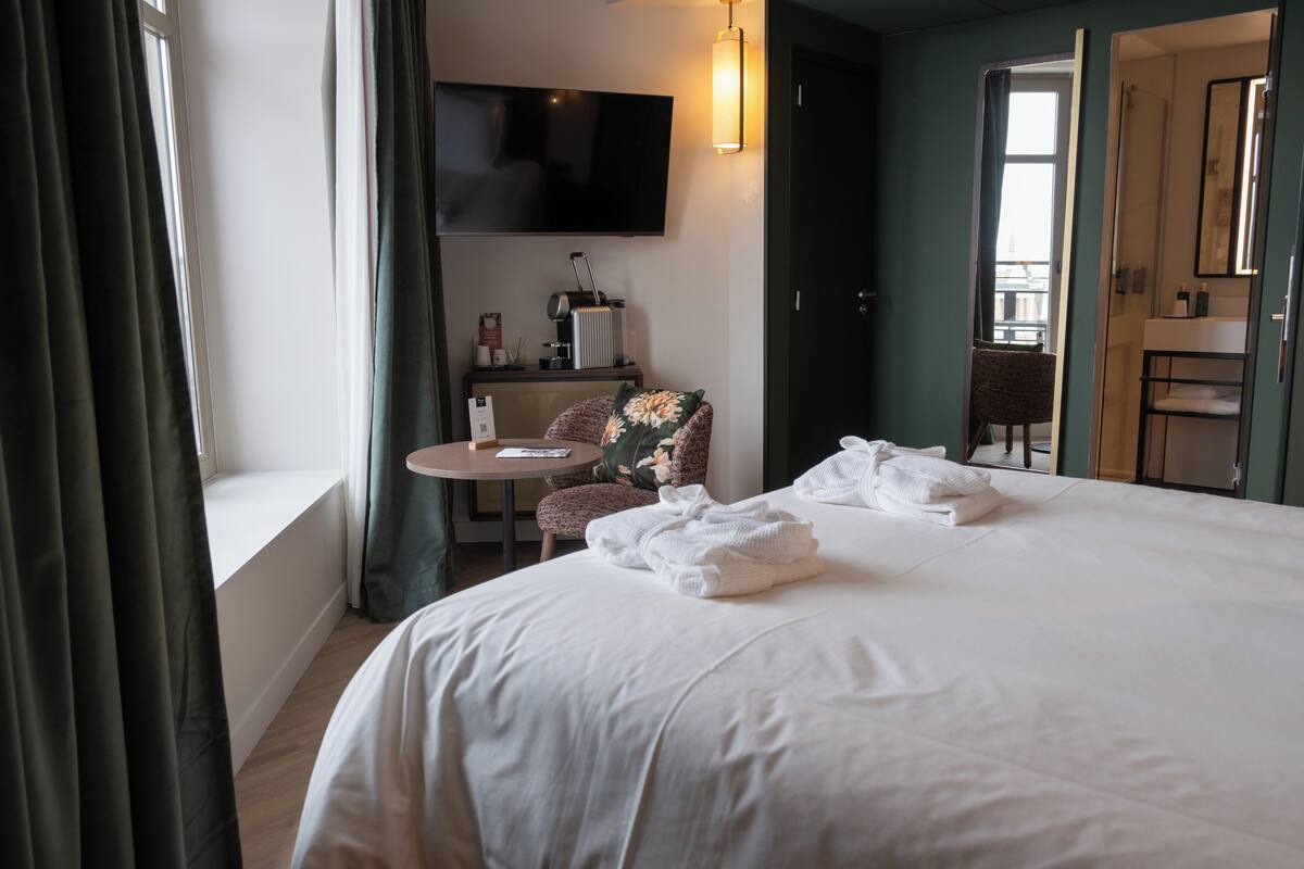 A hotel room with a bed, a round table, a chair, a TV, and a bathroom visible through an open door.