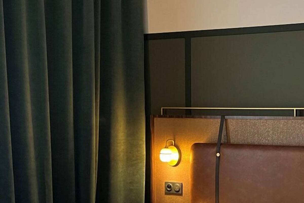 A room with a bed, a headboard with a light, and a curtain.