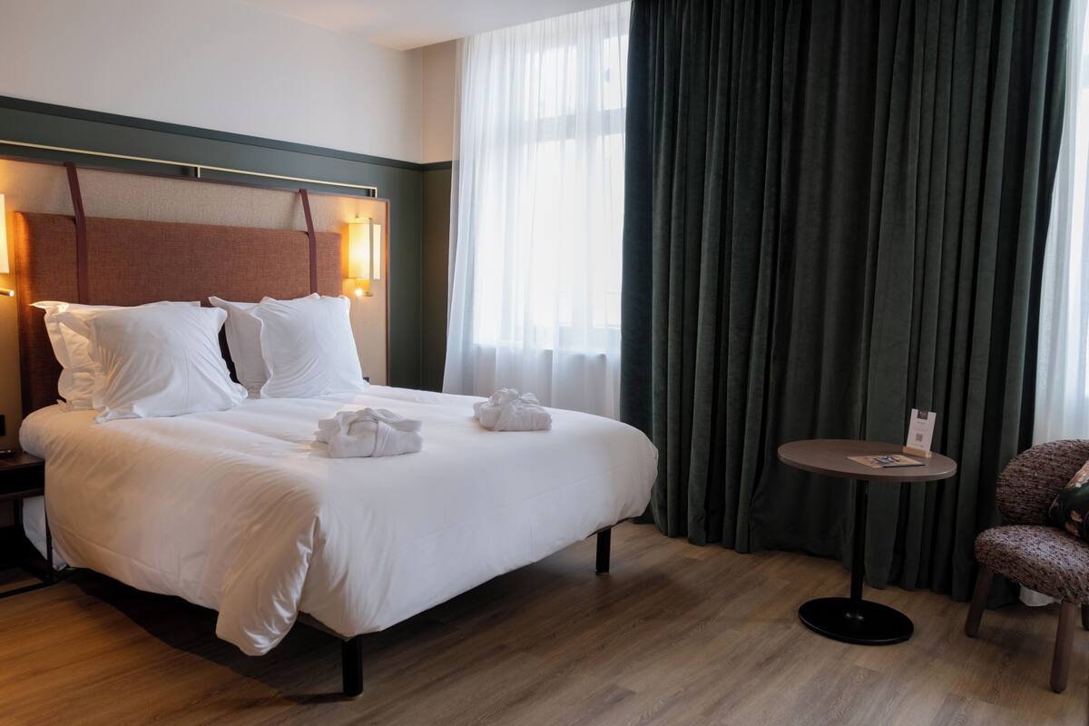 A hotel room with a large bed, a round table, a chair, and a window with curtains.