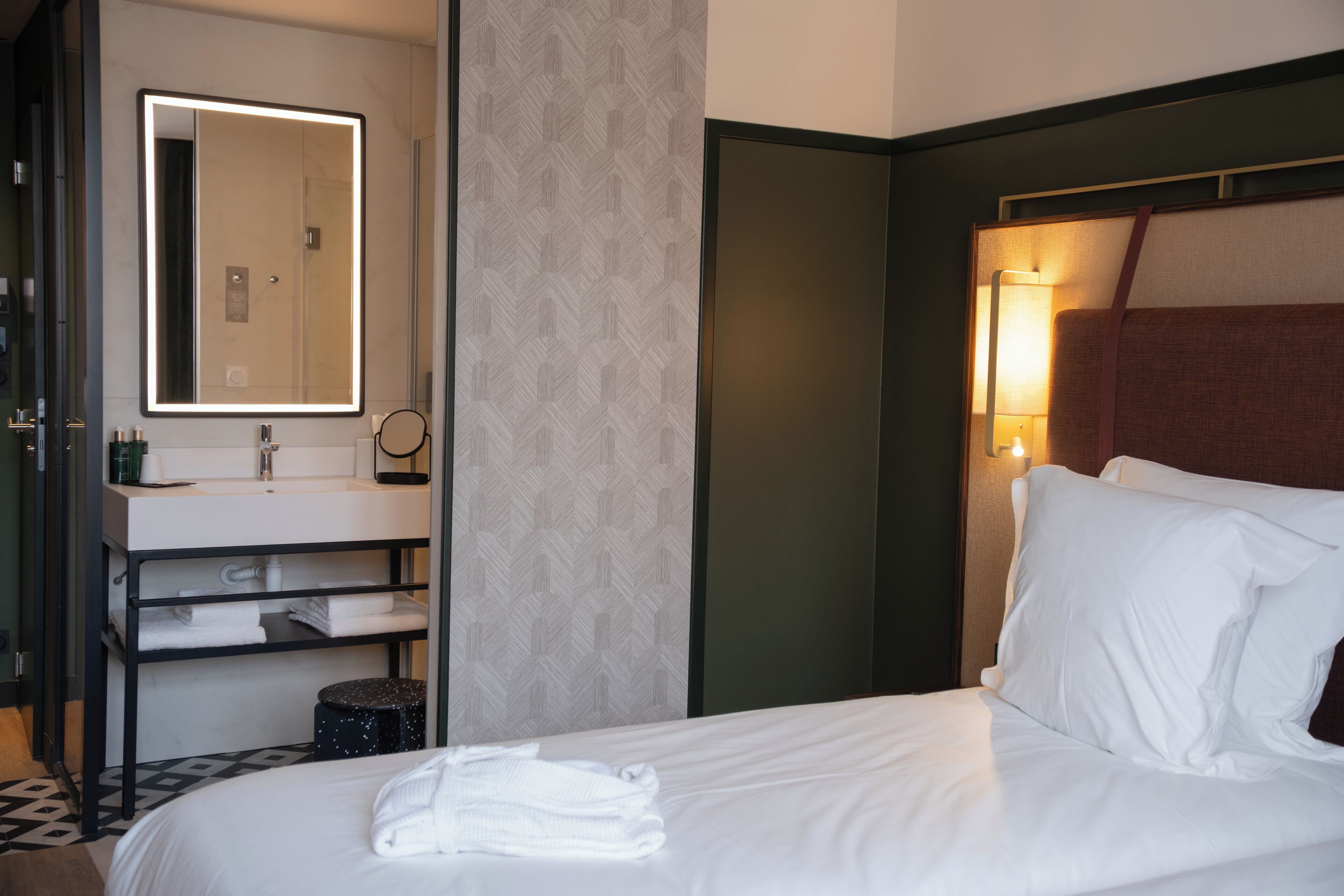 Hotelkamer met een bed, nachtkastje, spiegel, wandlamp en badkamer met wastafel en handdoekhouder.